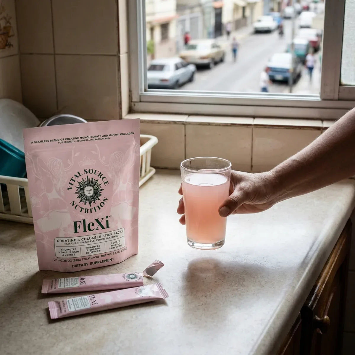Vaso con bebida rosada de creatina sabor piña junto a su sobre y su bolsa en un mesón de cocina